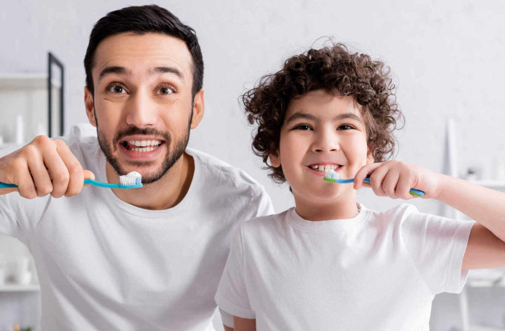 A father and son in white shirts and both holding a toothbrush with toothpaste on it and about to brush their teeth.