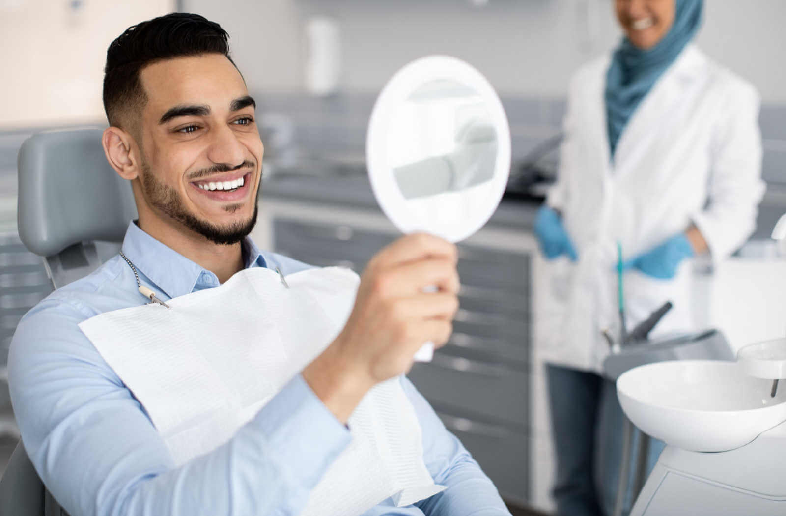 A man sitting on a dental chair with a mirror in his hand and checking his new veneers with a big smile on his face.